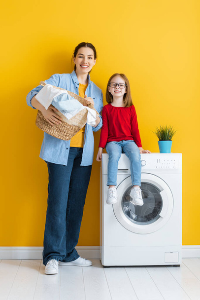 Beautiful young woman and child girl little helper are having fun and smiling while doing laundry at home. - Фото, изображение