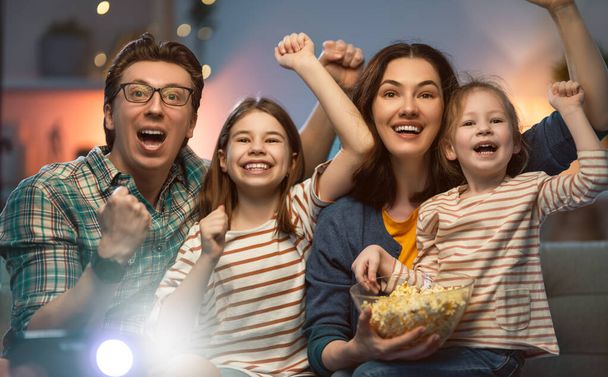 Happy family watching projector, TV, movies with popcorn in the evening at home. Mother, father and daughters spending time together. - Foto, Imagem