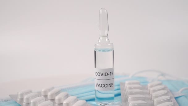 Ampul met een heldere vloeistof genaamd COVID-19 VACCINE op blauwe beschermende medische maskers. Met pakjes pillen. De camera gaat uit. - Video