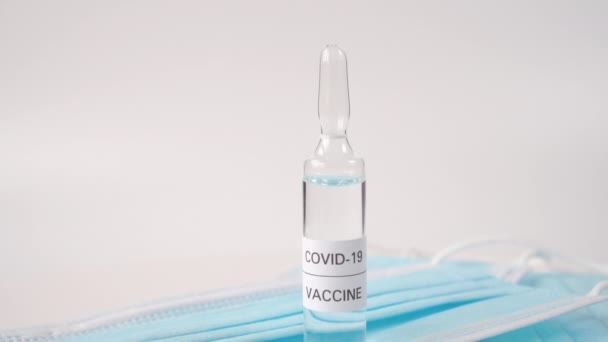 Ampola de vidro com um líquido transparente chamado COVID-19 VACCINE em máscaras médicas de proteção azul. Numa superfície branca. A câmara desce.
 - Filmagem, Vídeo
