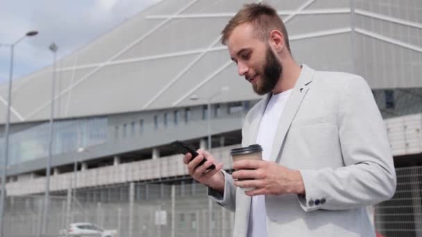 Ένας νεαρός επιχειρηματίας με αυτοπεποίθηση με γενειάδα κάθεται στις σκάλες και κοιτάζει ένα smartphone πίνοντας καφέ στο παρασκήνιο του σύγχρονου κτιρίου. Καφέ break.Light κομψό κοστούμι, παγκόσμιο δίκτυο. - Πλάνα, βίντεο