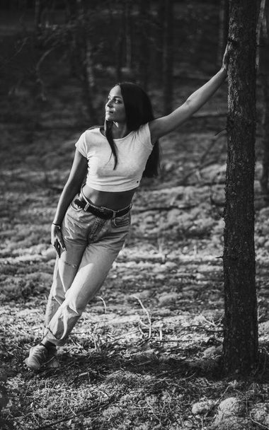 Young trendy pretty woman in moms jeans and a white top is leaning and holding her hand on a tree in the summer pine forest. Black and white photo - Photo, Image