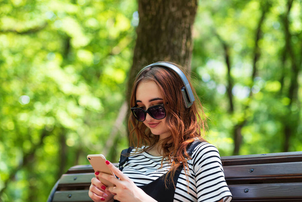 A young pretty red haired girl is listening to music with headphones while sitting on a Park bench - Foto, immagini