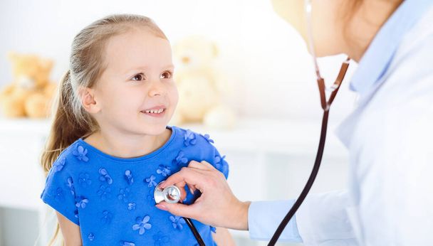 Doctor examining a child by stethoscope in sunny clinic. Happy smiling girl patient dressed in blue dress is at usual medical inspection - Foto, imagen