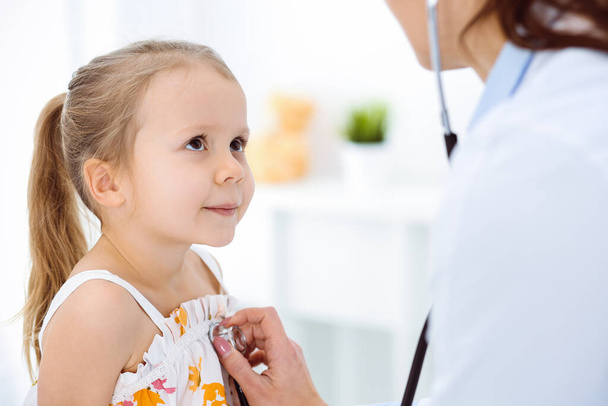 Doctor examining a child by stethoscope in sunny clinic. Happy smiling girl patient dressed in bright color dress is at usual medical inspection - Foto, imagen