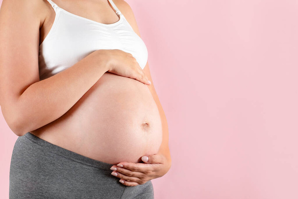 Close up of pregnant woman wearing supportive seamless maternity bra & grey yoga pants, arms on her belly. Female hands wrapped around big bare tummy. Child expectancy concept. Background, copy space - Photo, image
