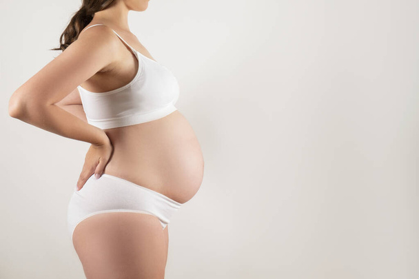 Close up of pregnant woman wearing supportive seamless maternity bra and maxi bottoms. Female hands wrapped around big bare tummy. Child expectancy concept. Background, copy space - Photo, Image