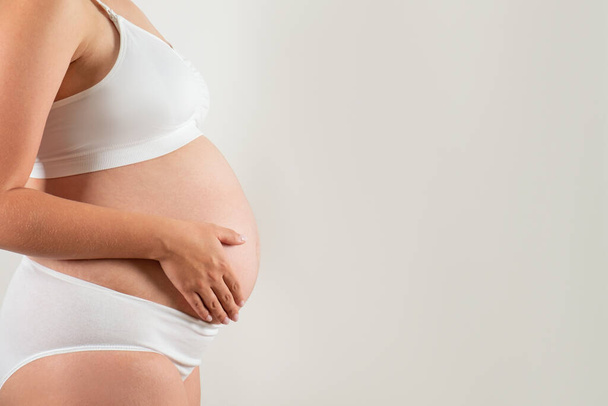 Close up of pregnant woman wearing supportive seamless maternity bra and maxi bottoms, arms on her belly. Female hands wrapped around big bare tummy. Child expectancy concept. Background, copy space - Foto, immagini