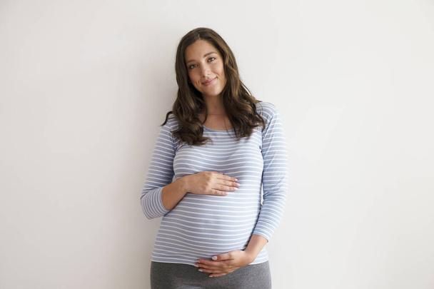 Portrait shot of young beautiful woman on third trimester of pregnancy. Close up of pregnant female with arms on her round belly. Expecting a child concept. Background, copy space. - Foto, imagen