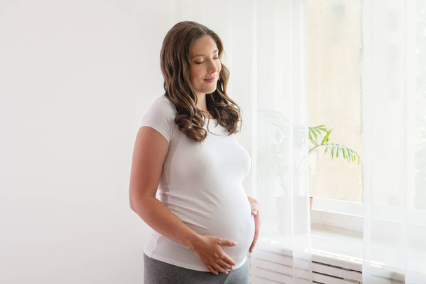 Portrait shot of young beautiful woman on third trimester of pregnancy. Close up of pregnant female with arms on her round belly. Expecting a child concept. Background, copy space. - Foto, afbeelding