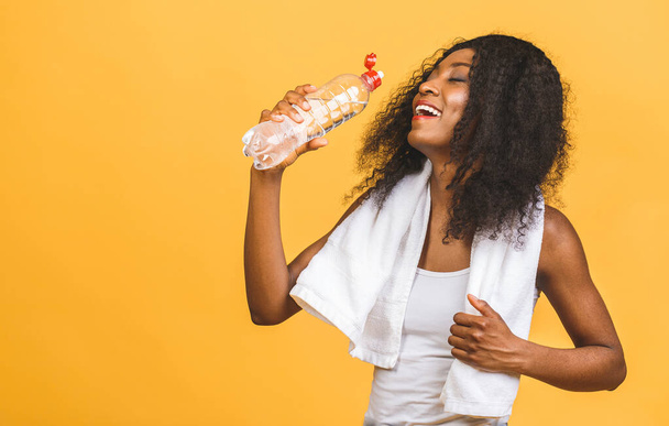 Όμορφη ελκυστική νεαρή ιδρωμένη Αφροαμερικανή μαύρη γυναίκα που πίνει νερό μετά την άσκηση προπόνηση, ενυδατώνει τη δίψα σβήνοντας, απομονώνεται σε κίτρινο φόντο. - Φωτογραφία, εικόνα