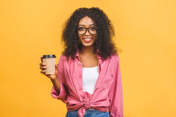 Portrait of african american woman 20s with afro hairdo looking aside while drinking takeaway coffee or tea from paper cup isolated over yellow background. - Photo, Image