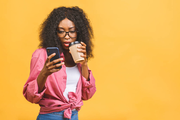 Feliz jovem afro-americana casualmente vestida de pé isolado sobre fundo amarelo, falando no celular, segurando takeaway xícara de café
. - Foto, Imagem