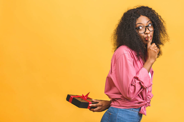 Happy african american lady in casual looking aside and laughing while holding present isolated over yellow background. - 写真・画像
