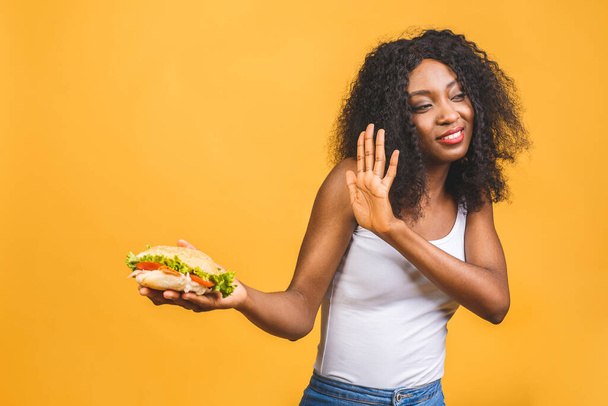Stop sign. Portrait of her she nice attractive lovely sad african american girl holding in hand tasty burger having gastritis isolated on yellow background. - 写真・画像