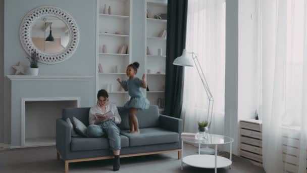Happy small African American preschool girl naughty daughter is jumping on sofa in living room while tired white mother tries to relax at home reading book. Concept of family, parenthood, togetherness - Кадри, відео