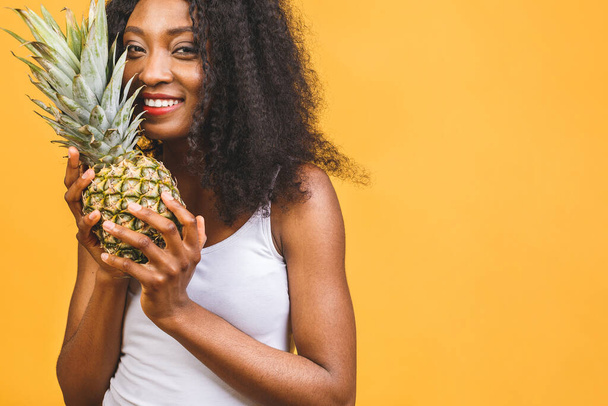 Ananasový exotický prostředník v dietě. Africká americká černoška s tmavou pletí veselá dívka drží ve svých rukou ananas izolované přes žluté pozadí.  - Fotografie, Obrázek