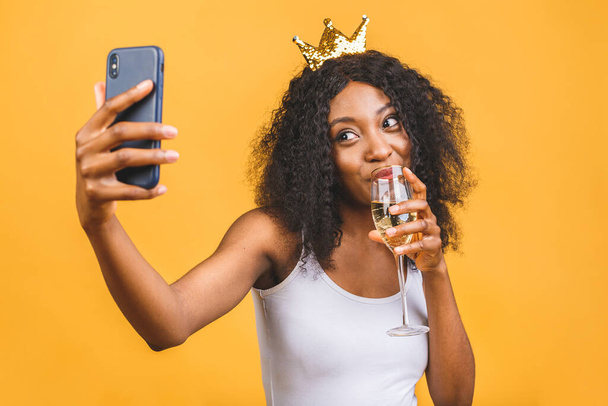 Feliz aniversário! Retrato de mulher negra afro-americana feliz com copo de champanhe e coroa dourada isolada sobre fundo amarelo. Usando telefone
. - Foto, Imagem