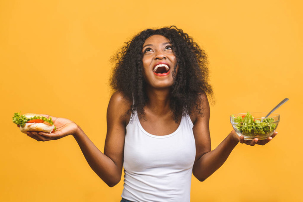 健康食品の概念。美しいアフリカ系アメリカ人黒人女性のクローズアップ黄色の背景に隔離されたサラダとバーガーを食べる.  - 写真・画像