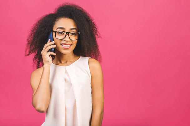 Retrato de una linda chica negra afro americana feliz en conversación casual en el teléfono móvil y riendo aislado sobre fondo rosa
. - Foto, Imagen