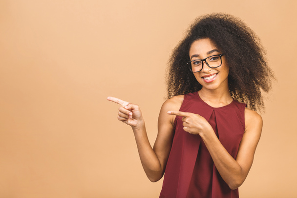 Retrato de una joven y feliz mujer rizada de negocios afroamericanos apuntando con los dedos al espacio de copia aislado sobre un fondo beige
. - Foto, imagen