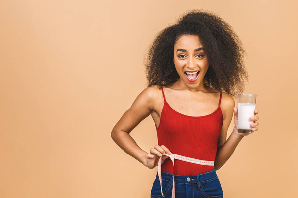 Ernährungskonzept. Schlanke Afroamerikanerin zeigt Zentimeter und hält Glas Joghurt isoliert über beigem Hintergrund.  - Foto, Bild