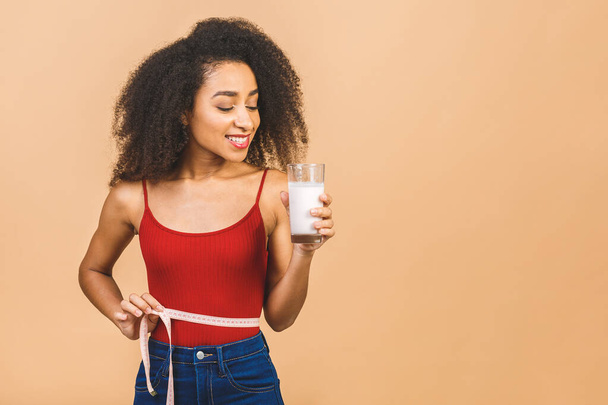Dieting concept. Slim african american woman shows centimeter and holding glass of yogurt  isolated over beige background.  - Foto, afbeelding