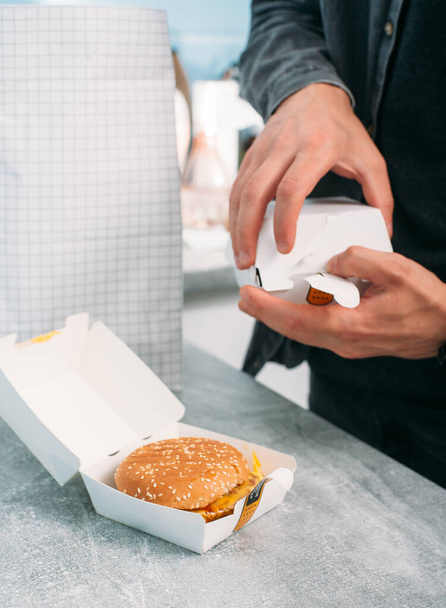 A guy in a gray shirt opens a paper box with a vegan burger. A food package with a food delivery in the background. - Foto, Imagem
