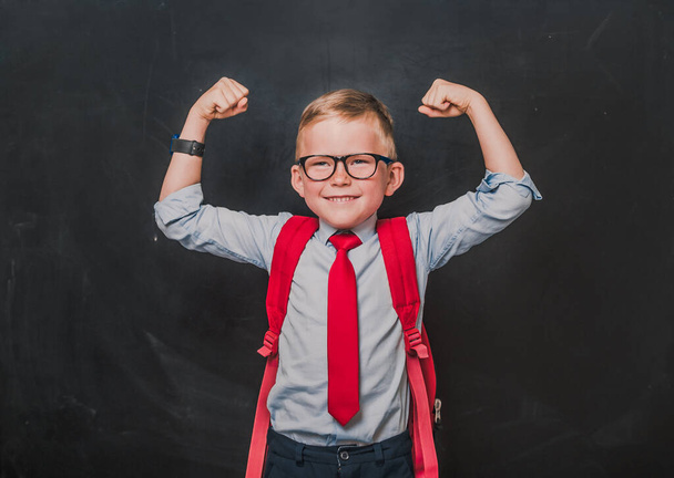 Education on fist class. Portrait of a schoolboy. Little pupil in uniform. Back to school. Funny strong kid in glasses and with bag against blackboard - Foto, Imagen
