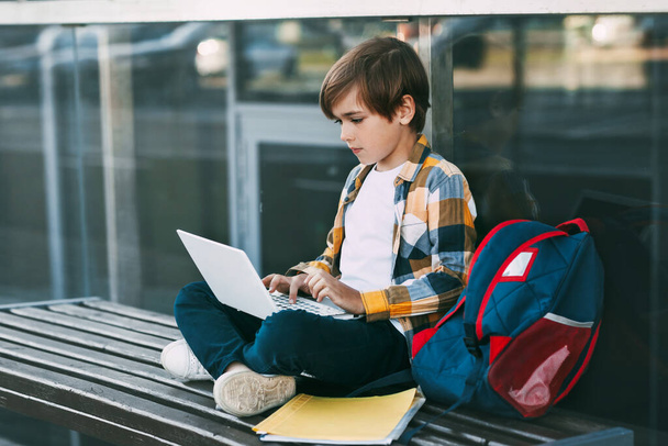 Um menino bonito em uma camisa xadrez está sentado em um banco com um laptop e digitando no teclado, ao lado de uma mochila
. - Foto, Imagem