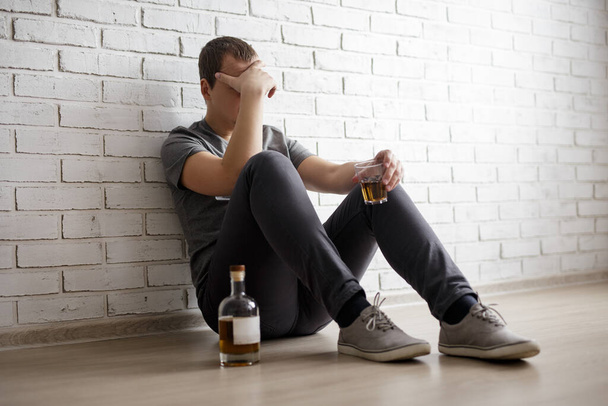 young man suffering from alcoholism, guy sitting on the floor with glass and whiskey bottle over white brick wall - Фото, изображение
