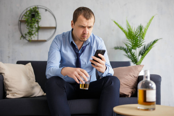 alcoholism, business and stress concept - tired businessman with smartphone drinking whiskey at home or office - Фото, изображение