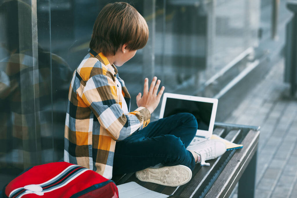 少年は、ベンチには、ラップトップとバックパックの横に座っている。少年はコンピュータを使って友達と通信し、手を振っている。教育、技術 - 写真・画像