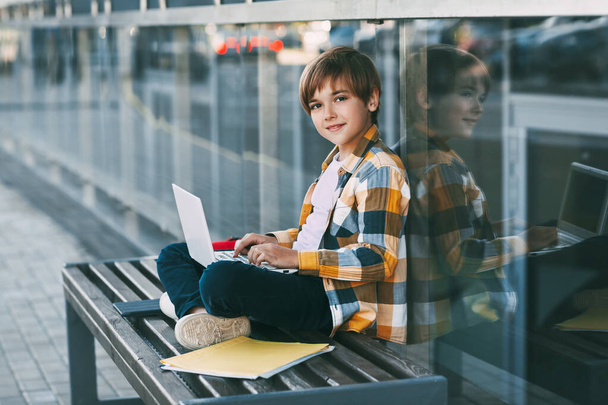 Un garçon mignon dans une chemise à carreaux est assis sur un banc avec un ordinateur portable et tapant sur le clavier, à côté d'un sac à dos. - Photo, image