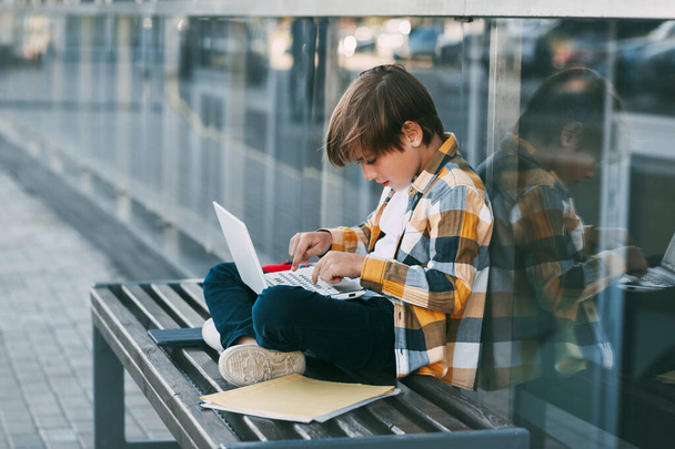 遊んだシャツを着たハンサムな男の子がベンチに座り、膝の上にノートパソコンを持っている。少年は友人とオンラインでテキストメッセージを送っている。社会的距離。教育、技術, - 写真・画像