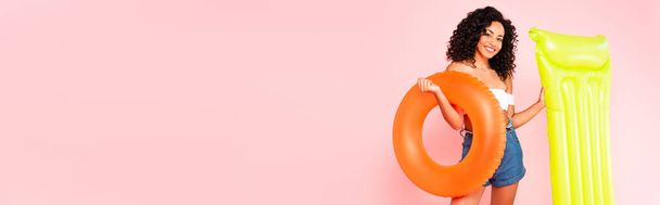 panoramic crop of cheerful african american girl holding swim ring and inflatable mattress isolated on pink  - Фото, изображение