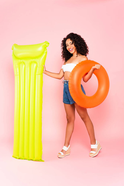 happy african american girl holding swim ring and inflatable mattress on pink  - Foto, imagen