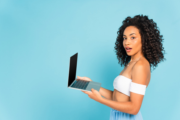 surprised and curly african american woman holding laptop isolated on blue - Fotó, kép