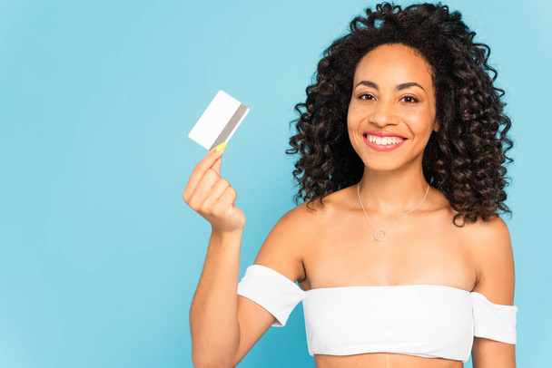 happy and curly african american woman holding credit card isolated on blue - Foto, imagen