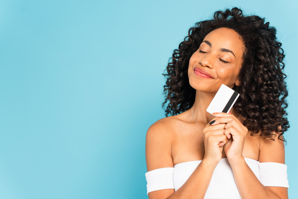 mujer americana africana feliz y rizada con los ojos cerrados sosteniendo la tarjeta de crédito aislada en azul
 - Foto, imagen