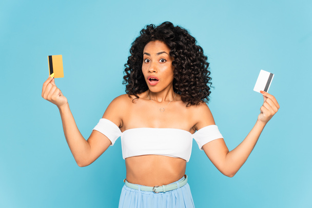 überrascht afrikanisch-amerikanische Frau hält Kreditkarten isoliert auf blau  - Foto, Bild
