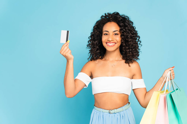 cheerful african american woman holding credit card and shopping bags isolated on blue  - Fotografie, Obrázek