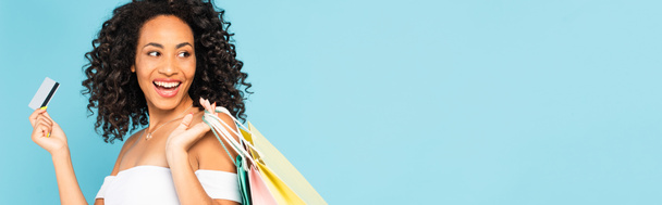 horizontales Bild einer aufgeregten afrikanisch-amerikanischen Frau mit Kreditkarte und Einkaufstaschen isoliert auf blauem Grund  - Foto, Bild