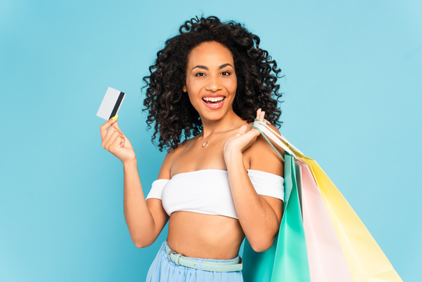 excited african american woman holding credit card and shopping bags isolated on blue  - Foto, Bild