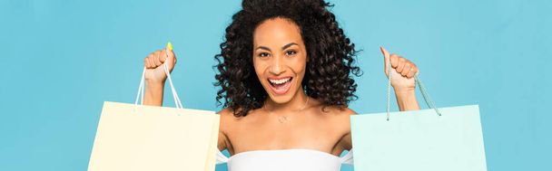 colpo panoramico di donna afro-americana eccitata che tiene borse della spesa e sorride isolato sul blu - Foto, immagini