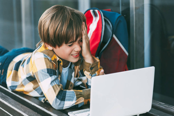 Ein fröhlicher Junge liegt auf einer Holzbank und arbeitet an einem Laptop neben einem Rucksack. Ein Schüler bereitet sich über das Internet auf den Schulunterricht vor. Soziale Distanz. Fernstudium, Ausbildung, Technologie - Foto, Bild