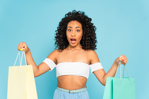 shocked african american girl with open mouth holding shopping bags isolated on blue - Фото, зображення