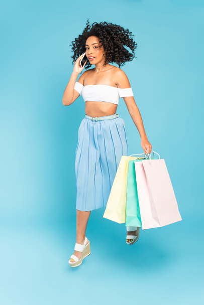 curly african american girl holding shopping bags and talking on smartphone on blue - Foto, imagen