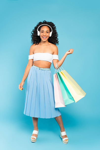 happy african american girl holding shopping bags and listening music in headphones on blue - Foto, Imagem