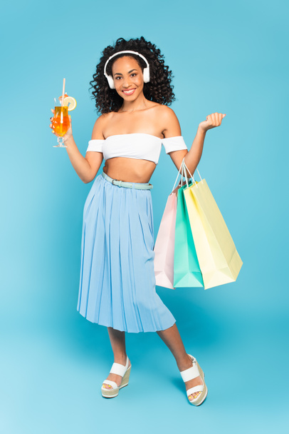 cheerful african american girl holding shopping bags and cocktail while listening music in headphones on blue - Zdjęcie, obraz